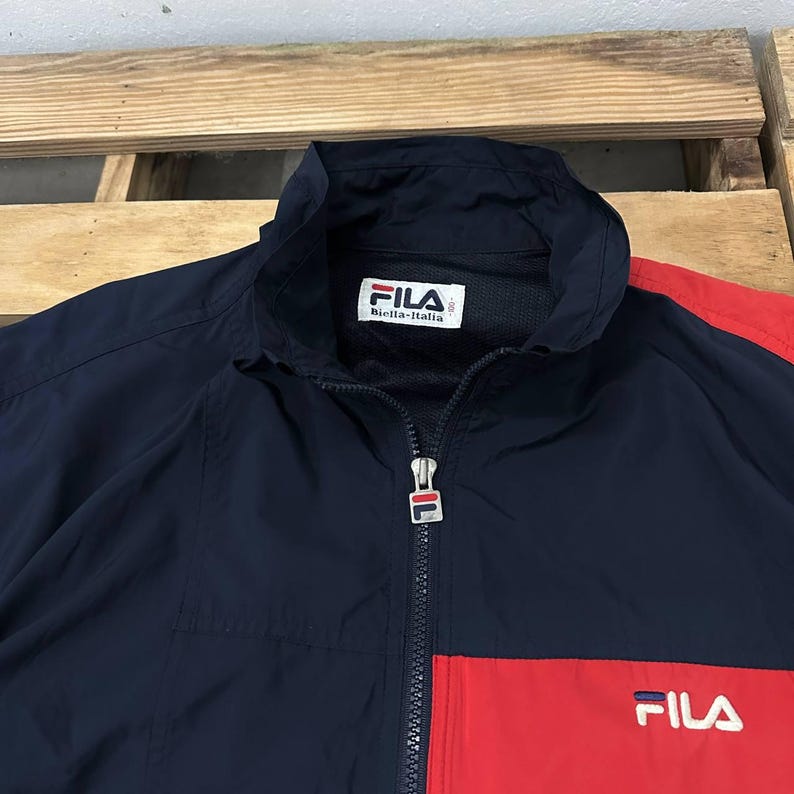 May include: A navy blue and red windbreaker jacket featuring a high collar, a full-length front zipper with a branded metal pull, and a white FILA Biella-Italia label at the inner neck. The front panel displays the FILA logo in white lettering.