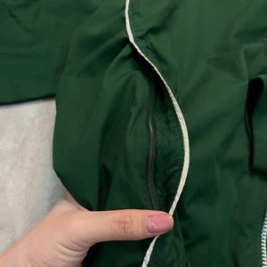May include: A dark green jacket with a white zipper and white trim. The jacket has a small white Nike logo on the upper left side. The jacket is made of a lightweight material.