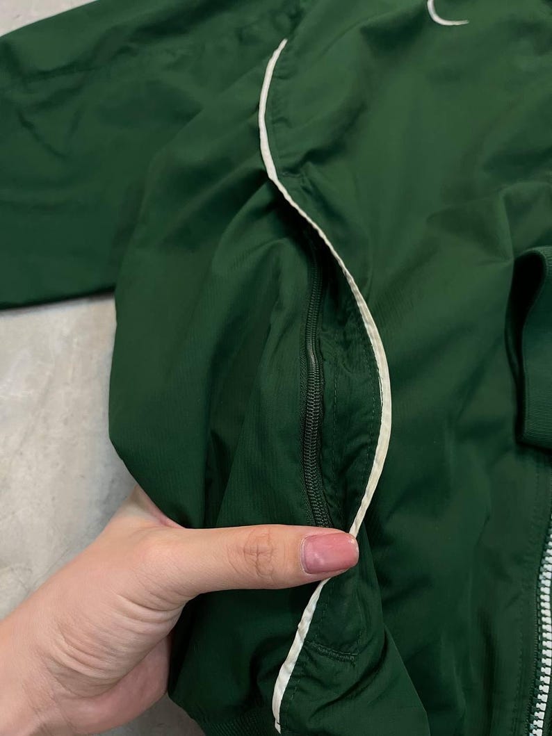 May include: A dark green jacket with a white zipper and white trim. The jacket has a small white Nike logo on the upper left side. The jacket is made of a lightweight material.