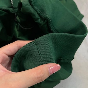 May include: Close-up of a dark green jacket sleeve. The sleeve features a ribbed cuff and a zipper. The fabric appears to be a smooth, possibly nylon or polyester material. The jacket is a solid, dark green color.