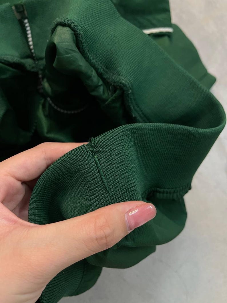 May include: Close-up of a dark green jacket sleeve. The sleeve features a ribbed cuff and a zipper. The fabric appears to be a smooth, possibly nylon or polyester material. The jacket is a solid, dark green color.