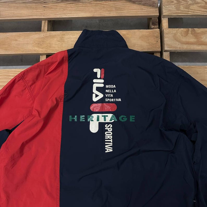 May include: A navy blue and red windbreaker jacket featuring a high collar and a large graphic on the back. The graphic displays the Fila logo in white and red, with the text "MODA NELLA VITA SPORTIVA" and the word "HERITAGE" in green letters.