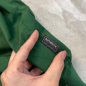 May include: Close-up of a dark green jacket sleeve. The sleeve has a ribbed cuff and a small rectangular tag that reads "AUTHENTIC TEAM APPAREL" in white text on a black background. The jacket appears to be made of a lightweight, possibly water-resistant material.