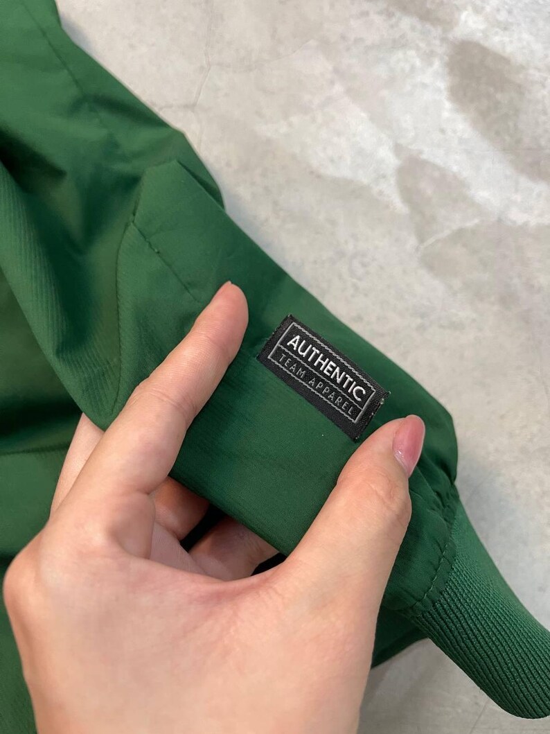 May include: Close-up of a dark green jacket sleeve. The sleeve has a ribbed cuff and a small rectangular tag that reads "AUTHENTIC TEAM APPAREL" in white text on a black background. The jacket appears to be made of a lightweight, possibly water-resistant material.