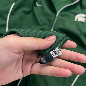 May include: Green athletic jacket with white accents and a white collar. Features a white Nike swoosh and a white logo on the right side. The jacket has a zipper closure and a ribbed knit cuff.