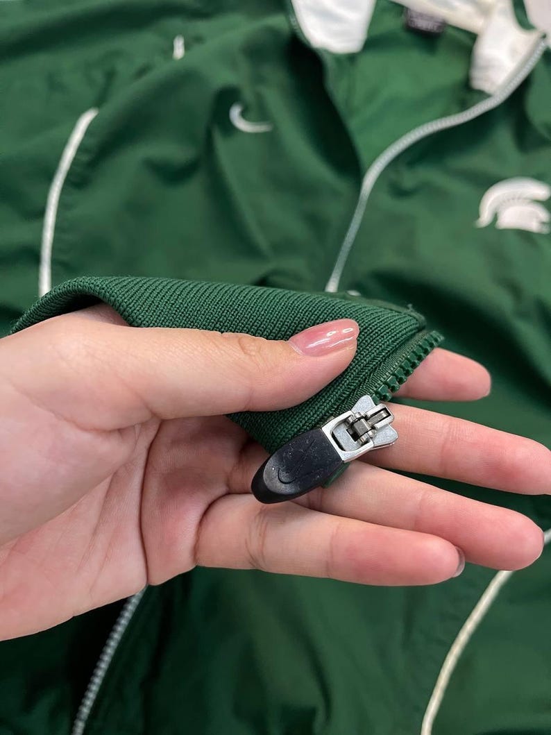 May include: Green athletic jacket with white accents and a white collar. Features a white Nike swoosh and a white logo on the right side. The jacket has a zipper closure and a ribbed knit cuff.