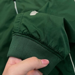 May include: A dark green jacket with a white zipper and a white logo on the chest. The jacket has a ribbed cuff and a white stripe down the side. The jacket is made of a lightweight material.