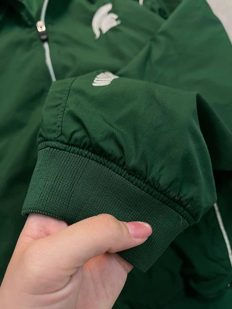 May include: A dark green jacket with a white zipper and a white logo on the chest. The jacket has a ribbed cuff and a white stripe down the side. The jacket is made of a lightweight material.