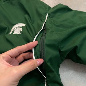May include: A dark green athletic jacket with a white Spartan helmet logo on the chest. The jacket has a white trim along the zipper and seams. The zipper is black. The jacket is made of a lightweight material.
