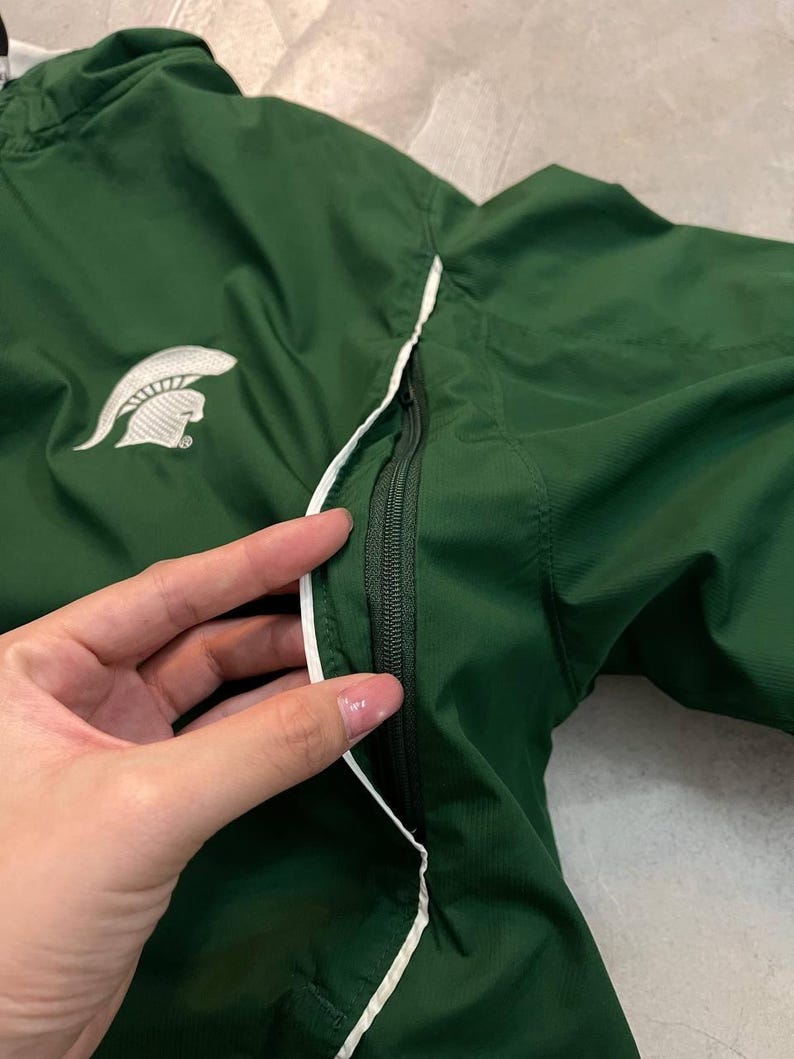 May include: A dark green athletic jacket with a white Spartan helmet logo on the chest. The jacket has a white trim along the zipper and seams. The zipper is black. The jacket is made of a lightweight material.