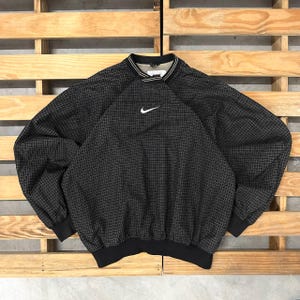 May include: A black long-sleeve pullover jacket featuring a subtle white grid pattern throughout. The garment has a mock neck collar with white striped detailing, a white Nike swoosh logo on the center chest, and solid black ribbed cuffs and hem.