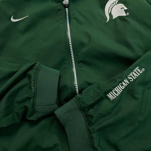 May include: Green athletic jacket with a white zipper, Nike logo, and the Michigan State Spartans logo. The sleeve cuff displays "MICHIGAN STATE" in white lettering. The jacket has a ribbed collar and cuffs.