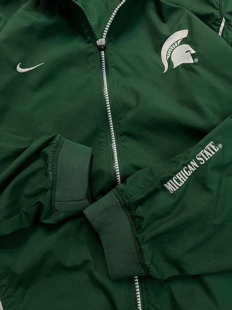 May include: Green athletic jacket with a white zipper, Nike logo, and the Michigan State Spartans logo. The sleeve cuff displays "MICHIGAN STATE" in white lettering. The jacket has a ribbed collar and cuffs.