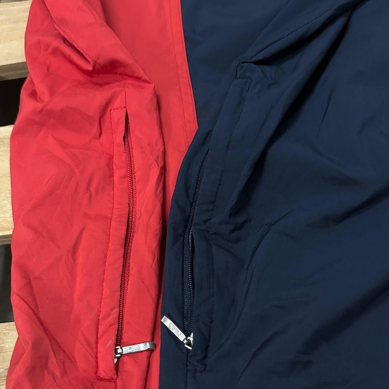 May include: A color-blocked jacket featuring a vibrant red section on the left and a deep navy blue section on the right. Each side includes a vertical zippered pocket with a silver-toned metal pull tab, providing functional storage for the garment.