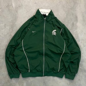 May include: Green athletic jacket with white trim and a white collar. Features a full zipper, the Nike swoosh, and a Spartan helmet logo. The jacket has a ribbed hem and cuffs.