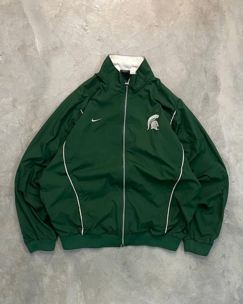 May include: Green athletic jacket with white trim and a white collar. Features a full zipper, the Nike swoosh, and a Spartan helmet logo. The jacket has a ribbed hem and cuffs.