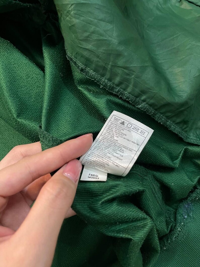 May include: Close-up of a dark green garment with a tag. The tag displays care instructions and the codes F6BSL and 14069X. The fabric has a textured, corduroy-like appearance.