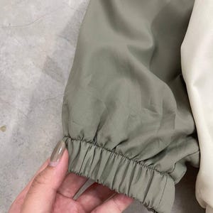 Peut inclure: Gros plan sur une manche de veste. La manche est vert olive avec un &eacute;lastique au poignet. La veste a un panneau blanc sur le c&ocirc;t&eacute; droit. Le tissu semble &ecirc;tre l&eacute;ger.