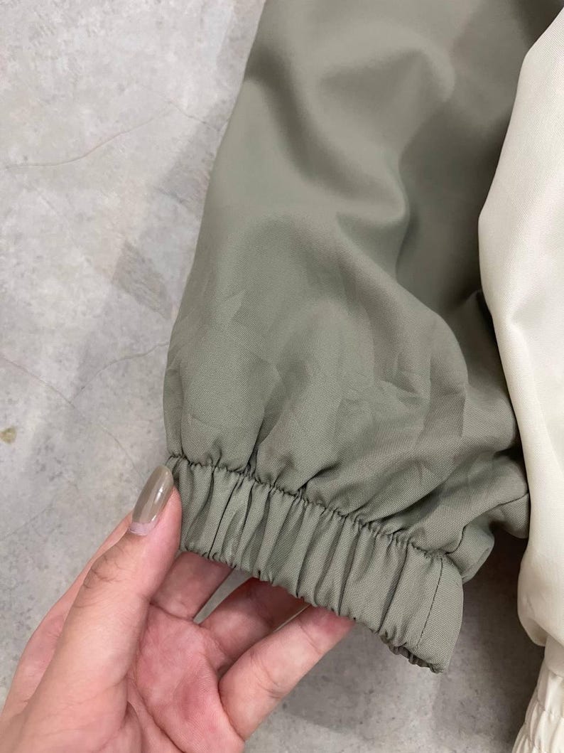 Peut inclure: Gros plan sur une manche de veste. La manche est vert olive avec un &eacute;lastique au poignet. La veste a un panneau blanc sur le c&ocirc;t&eacute; droit. Le tissu semble &ecirc;tre l&eacute;ger.