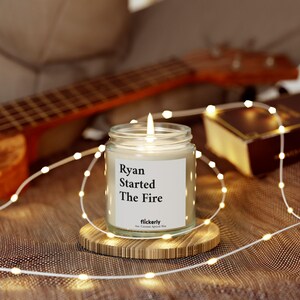 Puede incluir: Una vela blanca con una etiqueta negra que dice "Ryan Started The Fire" y "flickerly 100% Coconut Apricot Wax". La vela está encendida y se encuentra sobre un posavasos de madera rodeado de luces blancas.