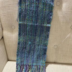 May include: A handwoven scarf in shades of blue, green, and purple. The scarf features a textured, striped pattern and long fringe at the bottom. The scarf is made of soft, lightweight material.