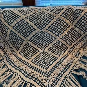 May include: A beige, handmade crochet shawl with a geometric pattern and fringe. The shawl is draped over a teal surface, showcasing its intricate openwork design and textured details. The fringe adds a decorative element to the edges.