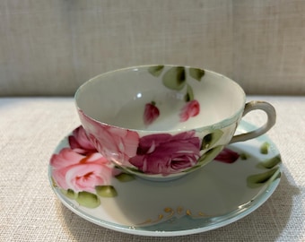 May include: A vintage teacup and saucer set. The cup and saucer are decorated with pink roses and green leaves on a light blue background. The cup has a small handle and the saucer has a gold trim.