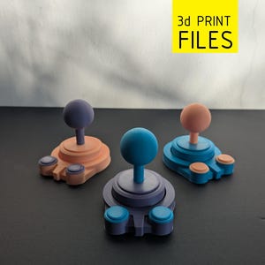 Puede incluir: Tres mandos de joystick impresos en 3D y coloridos. Cada mando tiene una base redonda con dos botones y un joystick central. Los joysticks son de color púrpura, azul y melocotón, con colores de base a juego. La imagen incluye el texto "3d PRINT FILES".