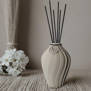 Könnte beinhalten: Eine beige, strukturierte Vase mit einem einzigartigen, geriffelten Design, die mehrere schwarze Diffusorstäbchen enthält. Ein kleines, rundes Etikett mit den Buchstaben "AH" ist angebracht. Die Vase ist Teil eines Raumduftsets.