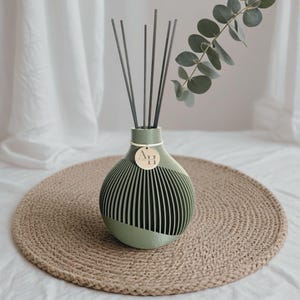 Könnte beinhalten: Ein grün-weißer dekorativer Diffusor mit schwarzen Stäbchen, platziert auf einer gewebten Jute-Matte. Der Diffusor hat ein einzigartiges Design mit vertikalen Linien und einem kleinen Holzanhänger. Eukalyptusblätter verleihen eine natürliche Note.