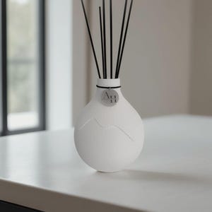 Könnte beinhalten: Ein weißer Keramikdiffusor mit schwarzen Stäbchen. Die Vase hat eine einzigartige, abgerundete Form mit einem bergähnlichen Design. Ein kleines, rundes Etikett mit dem Text "A11" ist angebracht. Der Diffusor steht auf einer weißen Oberfläche.