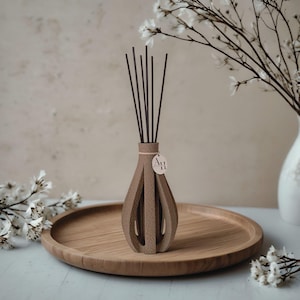 Könnte beinhalten: Ein brauner Diffusor-Vase mit offenem Design und dunklen Stäbchen, auf einem Holztablett. Die Vase hat ein Etikett mit der Aufschrift "Art 11". Weiße Blumen und eine weiße Vase sind im Hintergrund. Thema: Wohnkultur.