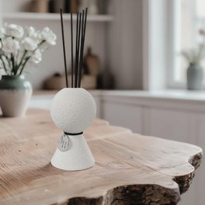 Könnte beinhalten: Ein weißer, strukturierter Keramik-Diffusor für ätherische Öle mit schwarzen Stäbchen. Der Diffusor hat eine kugelförmige Oberseite und einen konischen Sockel mit einem kleinen silbernen Anhänger mit der Aufschrift "AH 11". Der Diffusor steht auf einem Naturholztisch.
