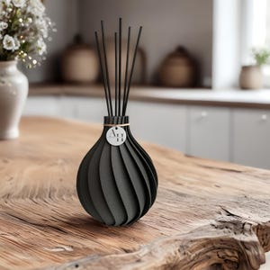 Könnte beinhalten: Ein schwarzer Reed-Diffusor mit einem einzigartigen, geschichteten Design. Der Diffusor hat eine runde Form mit vertikalen Rillen und einem kleinen, kreisförmigen Anhänger mit den Buchstaben "AH". Schwarze Stäbchen sind in die Oberseite des Diffusors eingesetzt.