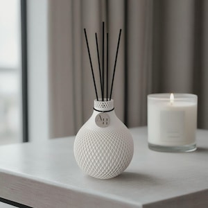 Könnte beinhalten: Ein weißer, kugelförmiger Diffusor mit Gittermuster, schwarzen Stäbchen und einem kleinen Anhänger. Rechts steht eine brennende Kerze in einem Glas. Der Diffusor und die Kerze stehen auf einer weißen Oberfläche.