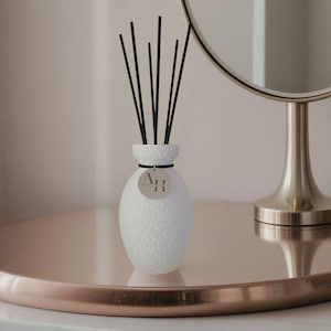 Könnte beinhalten: Ein weißer, strukturierter Keramikdiffusor mit schwarzen Stäbchen. Der Diffusor hat ein kleines, rundes Etikett mit den Buchstaben "A.II". Der Diffusor steht auf einem roségoldenen Tablett, mit einem silbernen Spiegel im Hintergrund.