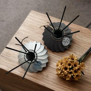 Könnte beinhalten: Zwei dekorative Schilf-Diffusoren mit schwarzen Schilfhalmen, einer silbernen und einer schwarzen, stehen auf einer Holzoberfläche. Jeder Diffusor hat ein kreisförmiges Design mit geschichteten Blütenblättern. Ein kleiner Strauß gelber Blumen ist ebenfalls vorhanden.