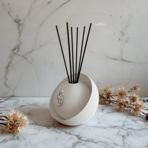 Könnte beinhalten: Ein weißer, strukturierter Keramik-Diffusor für ätherische Öle mit schwarzen Stäbchen. Der Diffusor hat ein einzigartiges kugelförmiges Design mit einem teilweisen Außenring. Ein kleines Etikett mit Text hängt am Diffusor. Getrocknete Blumen sind im Hintergrund.