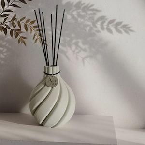 Könnte beinhalten: Ein hellgrüner Keramikdiffusor mit schwarzen Stäbchen. Die Vase hat ein Spiraldesign und ein kleines silbernes Etikett mit dem Text "Art 11". Der Diffusor steht auf einer weißen Oberfläche, mit einem Blattschatten im Hintergrund.