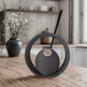 Könnte beinhalten: Ein dunkelgrauer Aromatherapie-Diffusor mit strukturierter Oberfläche und kreisförmigem Rahmen. Der Diffusor hat einen kleinen, runden Körper mit einem Etikett "A.H". Schwarze Stäbchen sind eingesetzt. Das Produkt steht auf einem Holztisch.