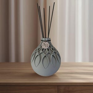Könnte beinhalten: Ein grauer dekorativer Diffusor mit einem gemusterten Design und schwarzen Stäbchen. Der Diffusor hat eine runde Form und ein Etikett mit den Buchstaben "AH". Der Diffusor steht auf einer Holzoberfläche.
