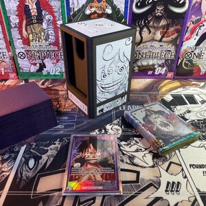 May include: A brown and black card game deck box with a black and white illustration of a character from the anime One Piece. The box is sitting on a table with a deck of cards and other One Piece card game products.