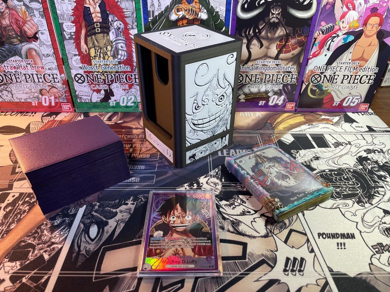 May include: A brown and black card game deck box with a black and white illustration of a character from the anime One Piece. The box is sitting on a table with a deck of cards and other One Piece card game products.