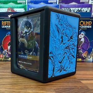 May include: Black card storage box with a clear front panel. The box features a blue embossed design on one side and holds a card labeled "Herald of the Arcane." The box is designed for trading card games.