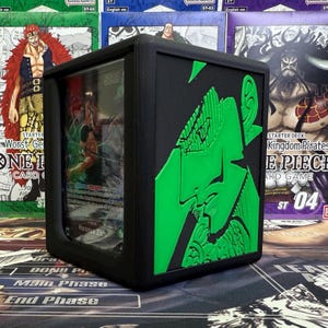 Puede incluir: Caja negra para guardar cartas con un diseño en relieve verde. La caja tiene un panel frontal transparente, que muestra una carta con el texto "5000" y "Roronoa Zoro". El fondo presenta varias cajas del juego de cartas "ONE PIECE".