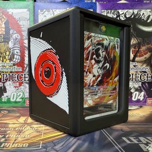 May include: Black card holder featuring a red and white eye design. It displays a card with a character illustration and the name "Sabo". Other card game boxes are visible in the background.