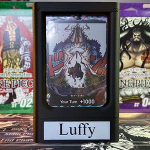 May include: A black card holder with a clear plastic window displaying a One Piece card featuring Luffy. The card has a blue and red design with the text "DOK!! CARD" and "Your Turn +1000". The card holder has a white label with the text "Luffy" on the front.