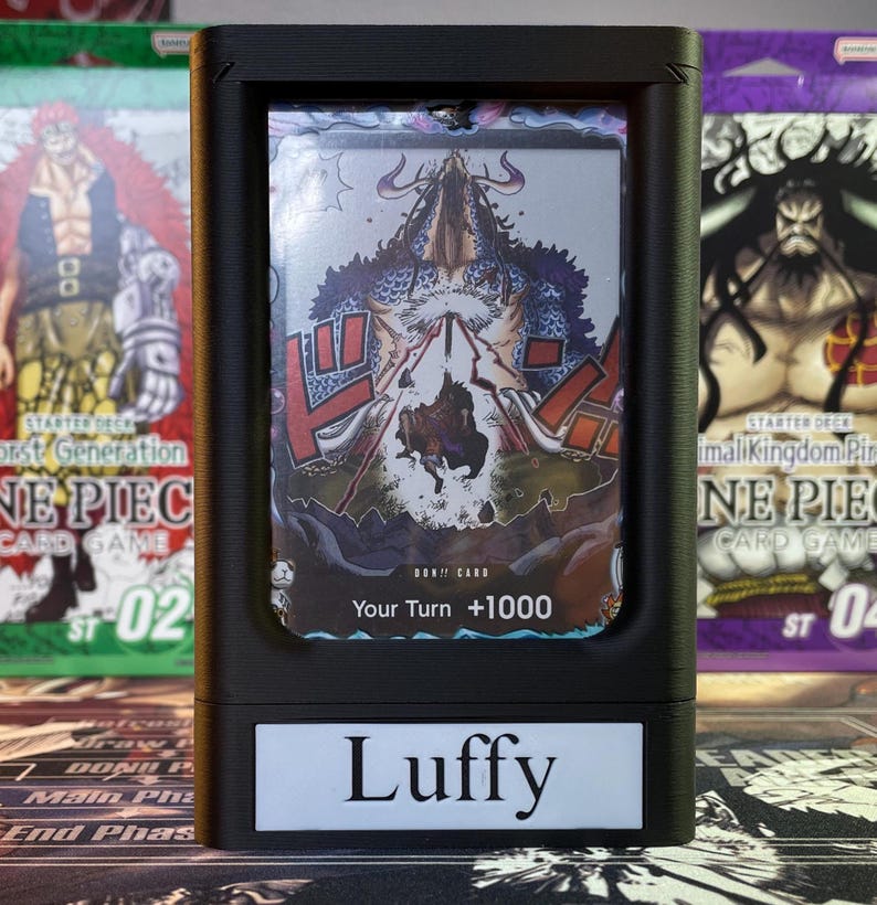 May include: A black card holder with a clear plastic window displaying a One Piece card featuring Luffy. The card has a blue and red design with the text "DOK!! CARD" and "Your Turn +1000". The card holder has a white label with the text "Luffy" on the front.