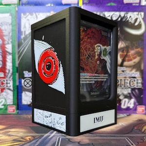 May include: Black card holder with a clear window displaying a trading card. The holder features a red and white eye design on the side and the text "IMU" on the front. The card shows a character with red and orange tones.