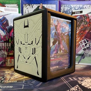 May include: A black and brown card storage box with a cream-colored embossed design of a Gundam robot on the front. The box is open, revealing a trading card with the text "Aile Strike Gundam". The box is surrounded by other Gundam card game boxes.